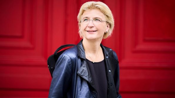 Frau mit schwarzer Lederjacke und Geigenkasten vor rotem Hintergrund 