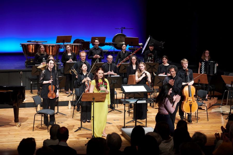 Eine Sängerin hält Rosen in der Hand und steht mit dem Ensemble Eisler Sinfonietta auf der Bühne im Studiosaal