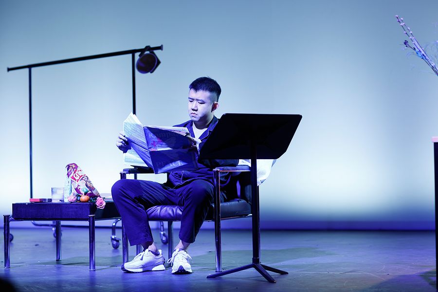Gesangstudent sitzt Zeitung lesend auf einem Sessel auf der bunt beleuchteten Bühne des Studiosaals