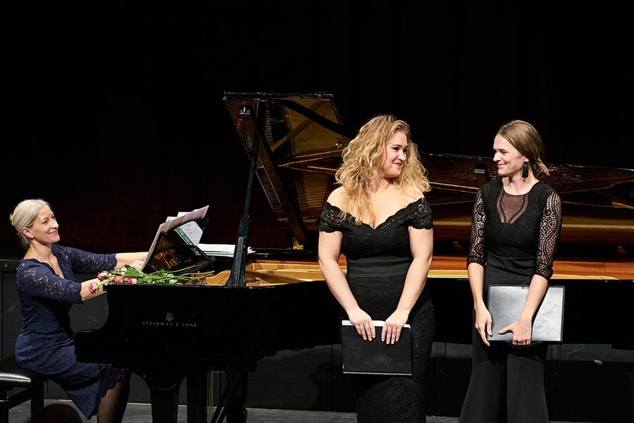 Zwei Studentinnen mit Notenmappen und Lehrende am Flügel auf einer Bühne