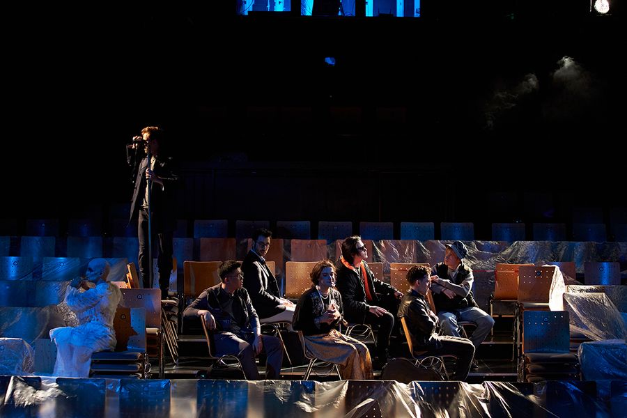 Sänger*innen sitzen auf mit Folien bedeckten Stühlen, einer steht mit Fernglas auf einem Stuhlstapel