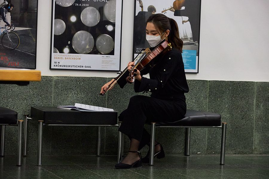 Studentin mit Mund-Nasen-Schutz übt ihre Streichpartie in einem Foyer, im Hintergrund Konzertplakate