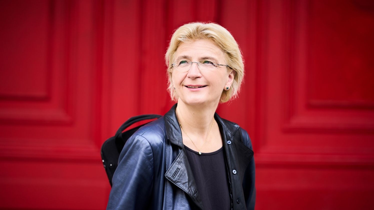 Frau mit schwarzer Lederjacke und Geigenkasten vor rotem Hintergrund 