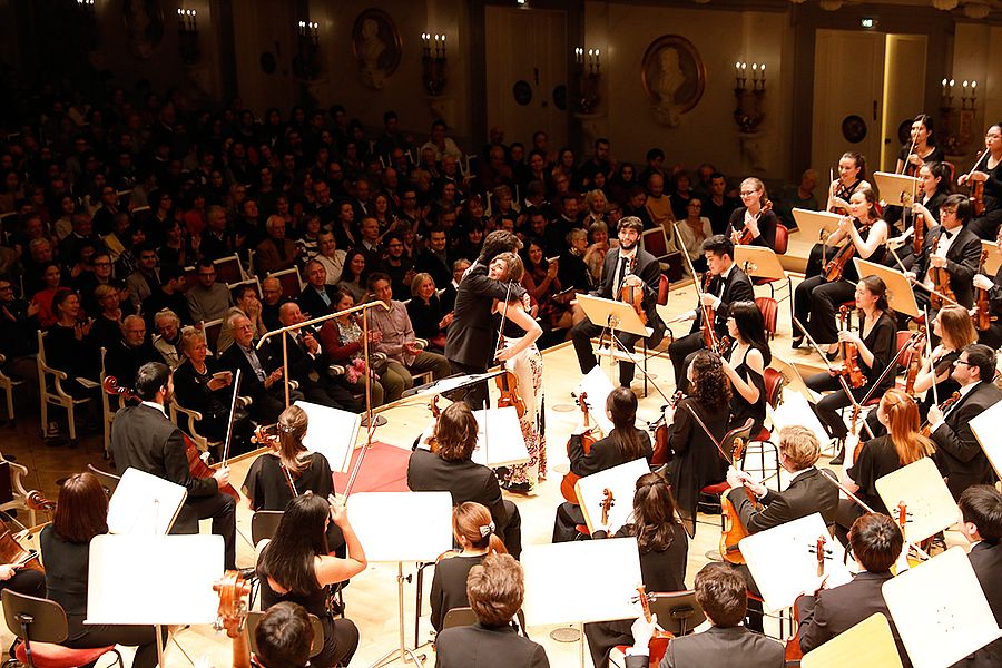 Dirigent und Geigensolistin umarmen sich vor dem Orchster, davor klatschendes Publikum