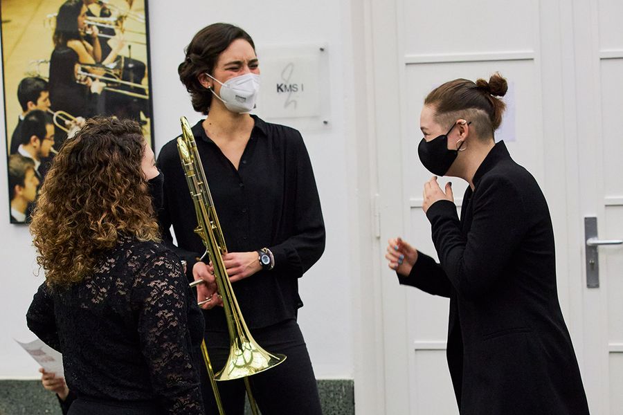 Drei Studentinnen, zwei davon mit Blasinstrumenten in der Hand, lachend und mit Mund-Nasen-Schutzwährend einer Konzertpause vor dem Saal