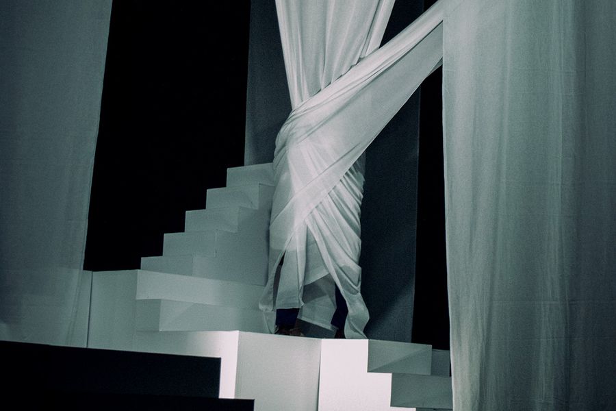Vollständig von einem weißen Vorhang eingehüllte Person in einem kontrastreich ausgeleuchteten Bühnenbild mit Treppen und langen Vorhängen