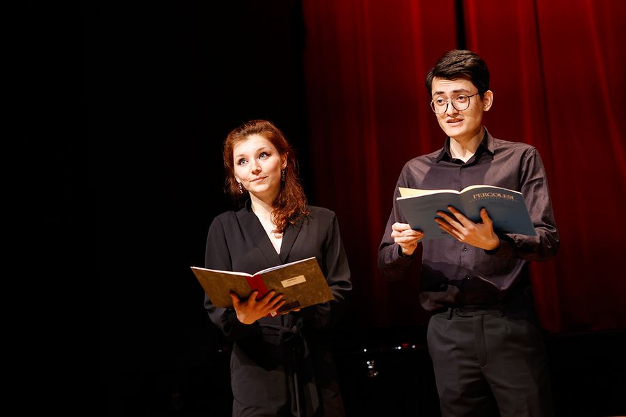 Gesangstudentin und Student halten Notenmappen in den Händen und singen auf der Bühne des Studiosaals vor rotem Vorhang
