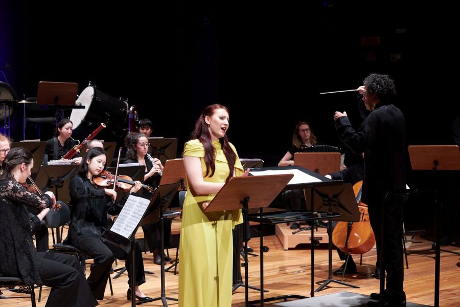 Eine Sängerin in einem gelben Kleid steht vor einem kleinen Orchester und singt