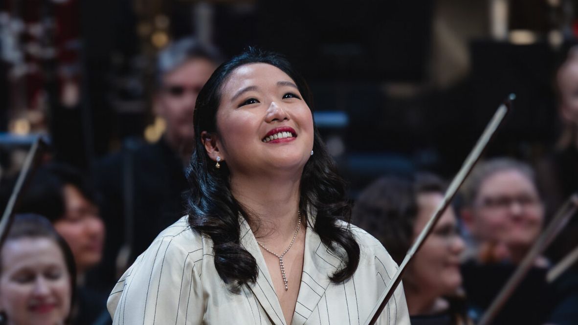 Geigerin verbeugt sich im Schlussapplaus vor dem Publikum mit Orchester im Hintergrund. 