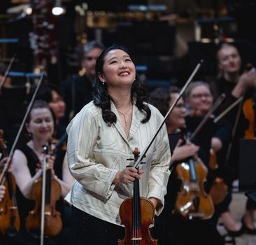 Geigerin verbeugt sich im Schlussapplaus vor dem Publikum mit Orchester im Hintergrund. 