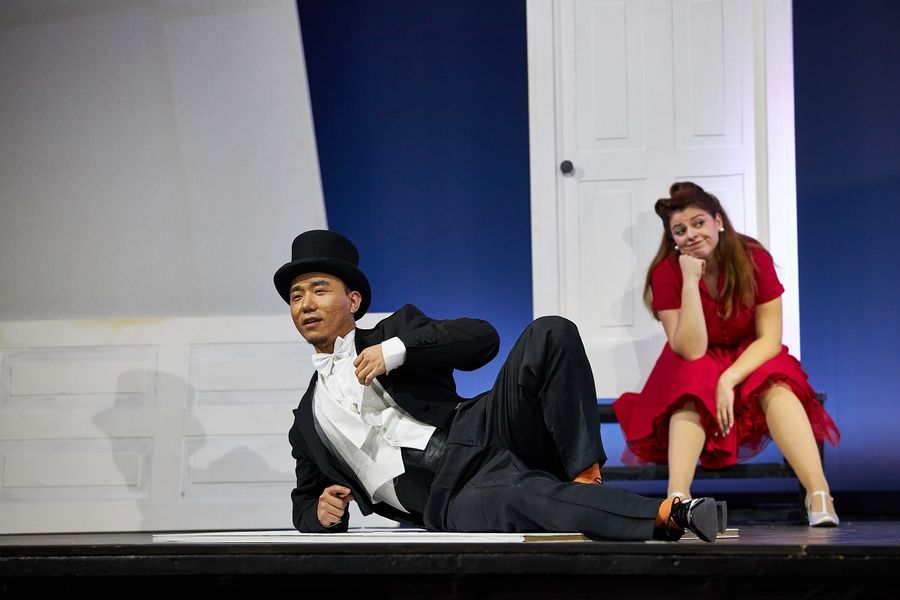 ein Sänger im Sakko liegt auf der Bühne, im Hintergrund eine Sängerin in einem roten Kleid