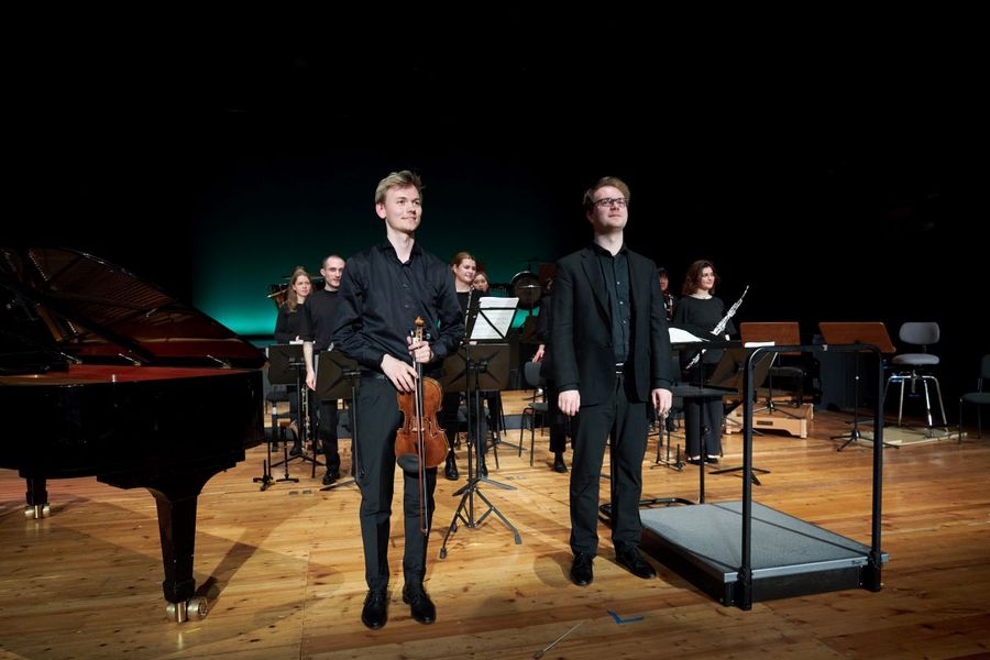 Zwei Männer, einer davon mit einer Geige in der Hand, stehen vor einem kleinen, ebenfalls stehenden Orchester