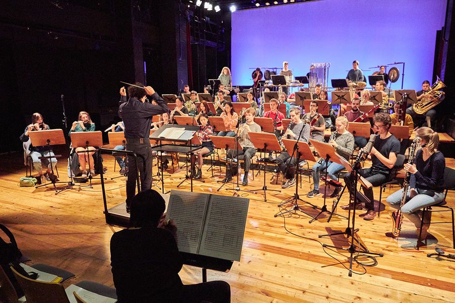 Professor Nawri dirigiert ein Blasorchester aus Studierenden auf der Bühne des Studiosaals während einer Probe