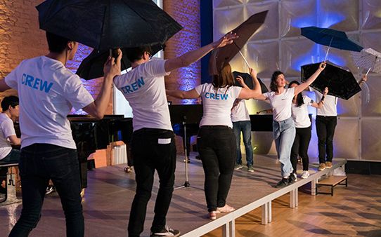 Studierende mit Regenschirmen auf einer Bühne vor einem Publikum mit Kindern