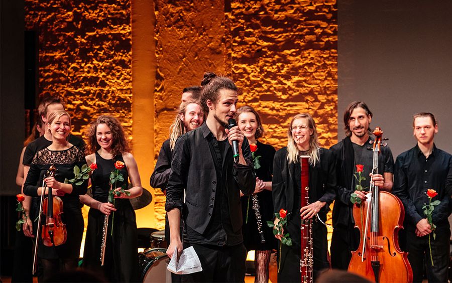 Leiter des Orchesters hält eine Rede, hinter ihm stehen Orchestermitglieder mit Instrumenten und Rosen