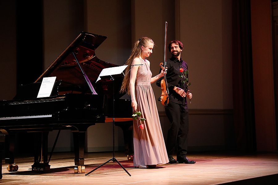 Studentin mit Geige und Rose und Pianist mit Rose stehen auf einer Bühne vor einem Flügel