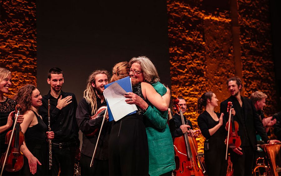 Die Fördervereinsvorsitzende umarmt ein Orchestermitglied, im Hintergrund weitere Orchestermitglieder mit Instrumenten