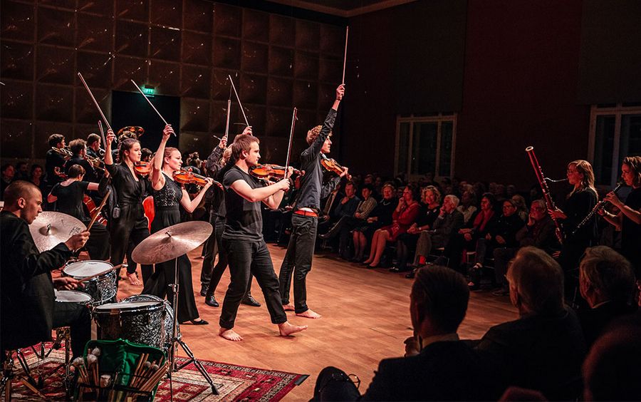 Personen mit Streichinstrumenten stehen mit erhobenen Bögen auf einer Bühne vor einem Publikum