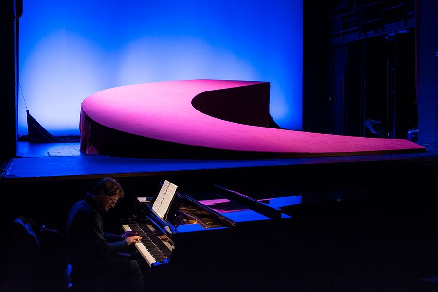 Im Vordergrund Pianist am Flügel sitzend, im Hintergrund grell ausgeleuchtetes Bühnenbild mit sichelförmiger Straßenlandschaft
