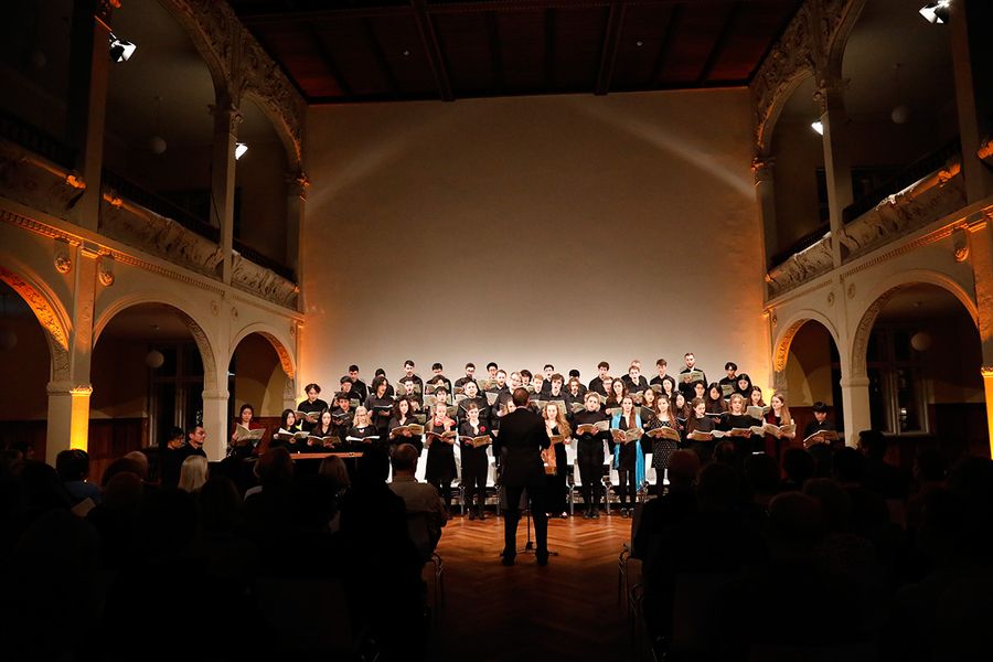 Chor aus Studierenden und Dirigent in einem Saal mit Publikum während eines Konzertes