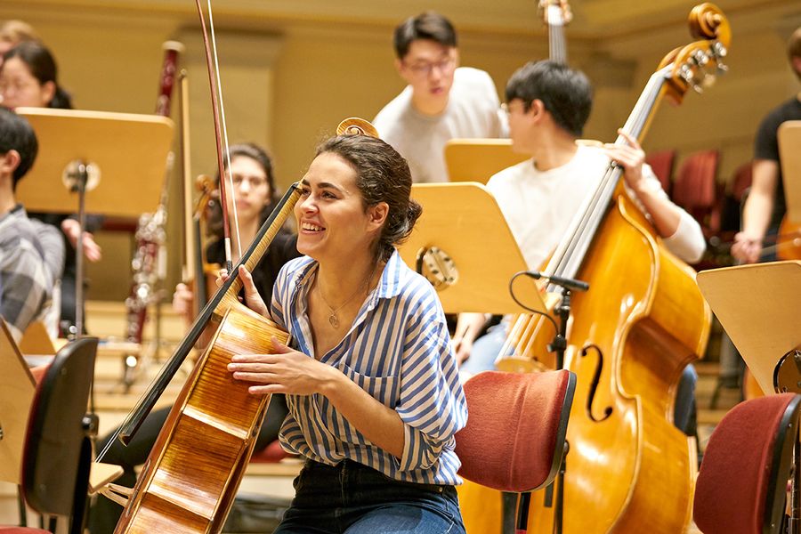 Studentin mit Cello inmitten von Studierenden mit Instrumenten während einer Probe