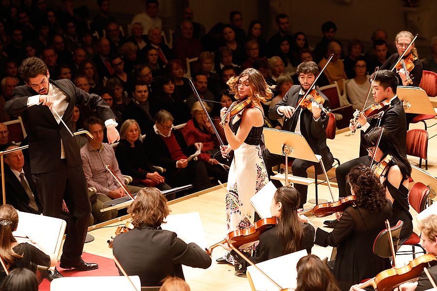 Dirigent, Geigensolistin und Streichersektion eines Orchesters vor Publikum