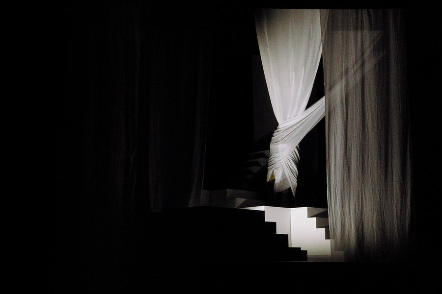 Vollständig von einem weißen Vorhang eingehüllte Person in einem kontrastreich ausgeleuchteten Bühnenbild in Schwarz-Weiß