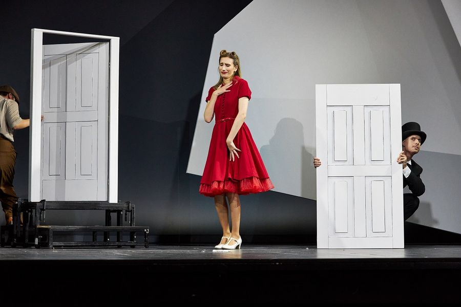 Sängerin in rotem Kleid steht auf der Bühne, neben ihr schaut hinter einer großen Tür ein Sänger vor