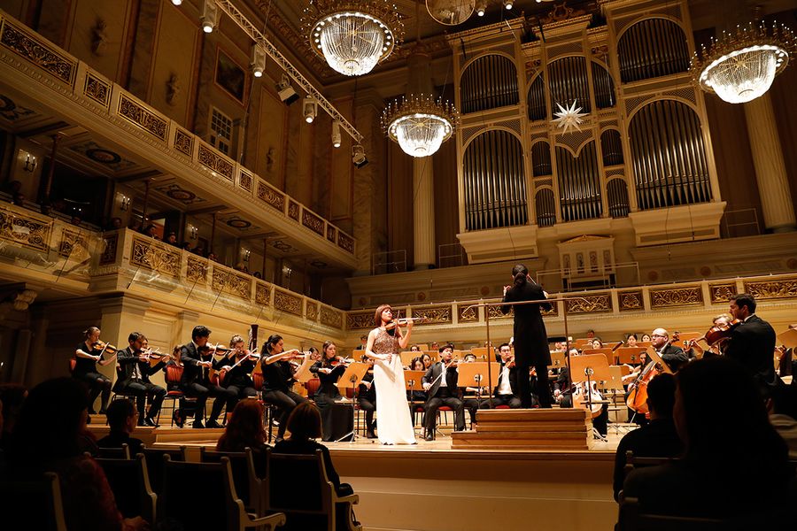 Orchester mit Geigensolistin auf einer Bühne in einem Saal mit Publikum während eines Konzertes