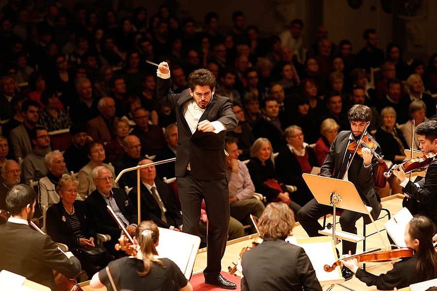 Publikum hinter einem Dirigent mit Taktstock der ein Orchester dirigiert