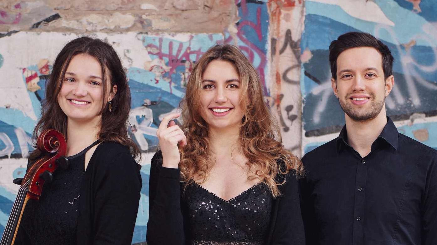 Zwei Musikerinnen und ein Musiker mit Instrumenten vor einer Graffiti-Wand