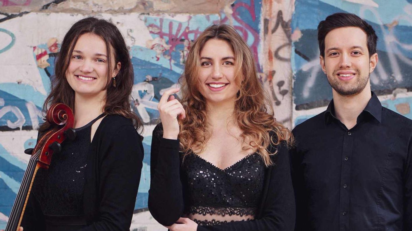 Zwei Musikerinnen und ein Musiker mit Instrumenten vor einer Graffiti-Wand