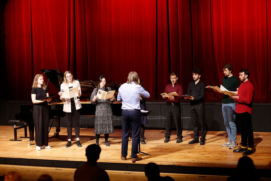 Gesangstudierende stehen singend mit Notenmappen auf der Bühne des Studiosaals, vor ihnen ein Dirigent