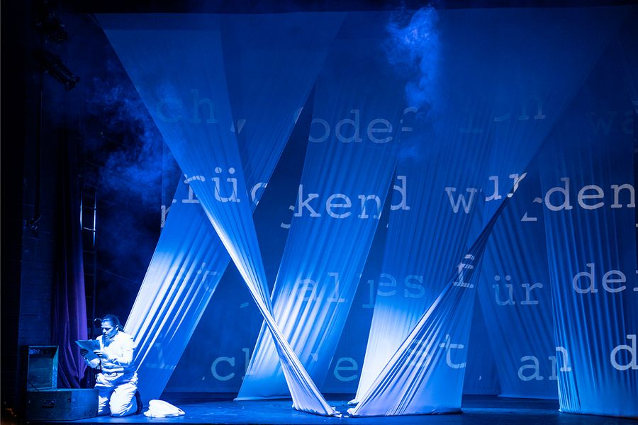 Gesangstudentin sitzt auf einer blau beleuchteten Bühne mit weißen von der Decke hängenden Tuchbahnen auf denen Worte geschrieben stehen