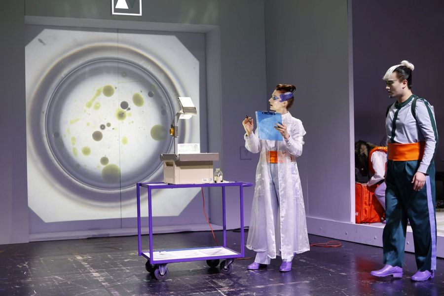 Ein Frau steht neben einem Overheadprojektor, im Hintergrund ein Bild von einem großen Reagenzglas