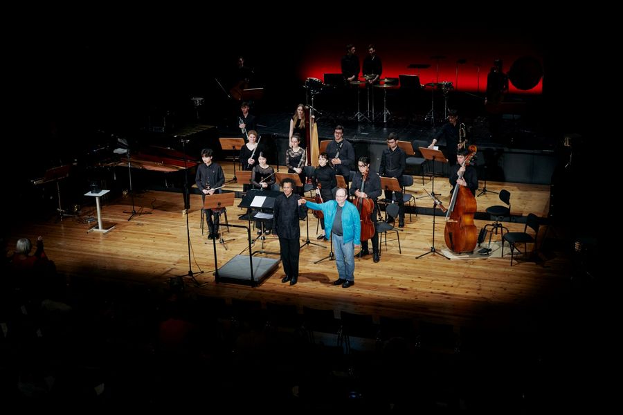 Ensemble, Dirigent und Mann in blauer Jacke nehmen auf der Bühne den Applaus stehend in Empfang