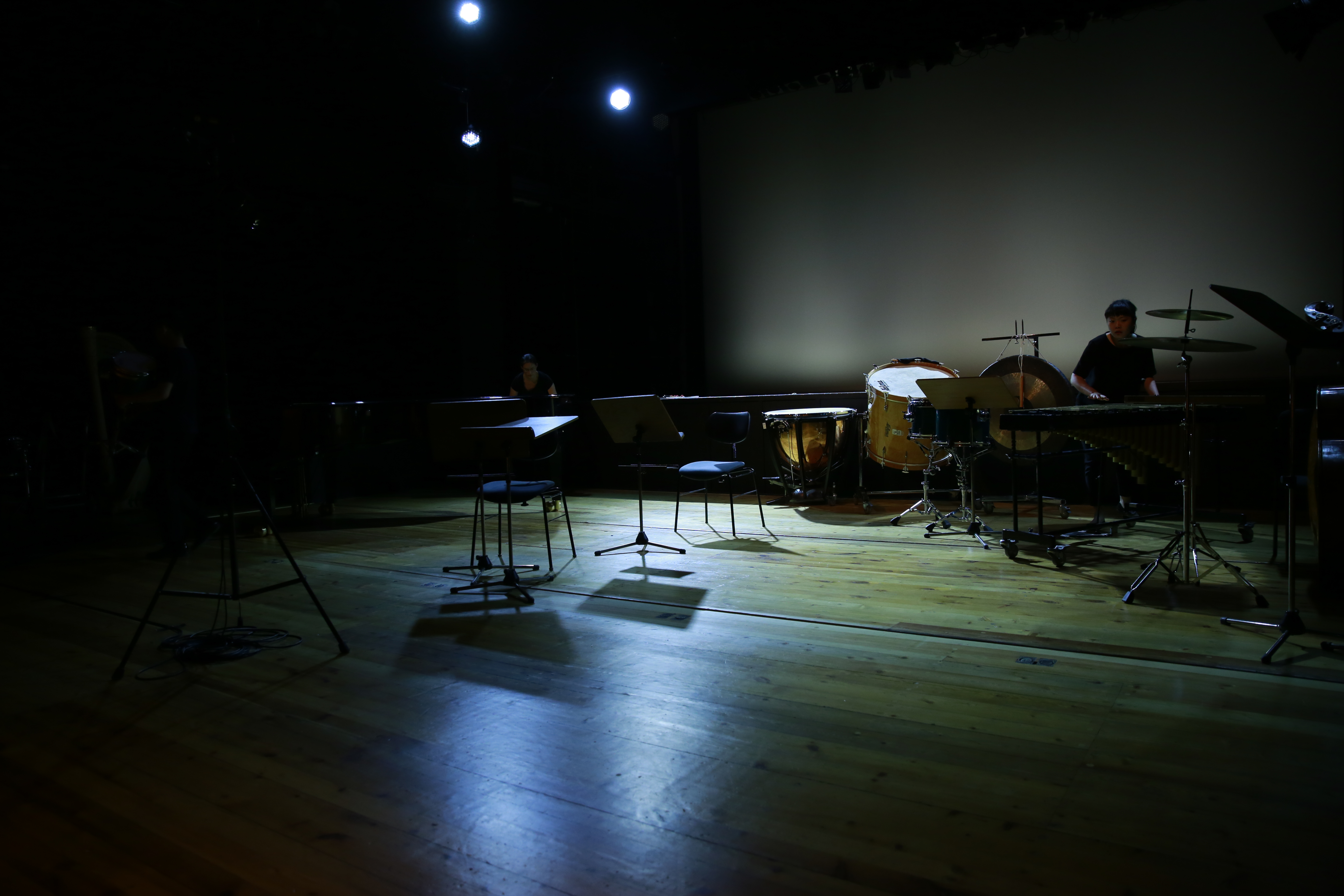 Dunkle Bühne mit Notenständern und Stühlen, dahinter eine Schlagzeug spielende Person