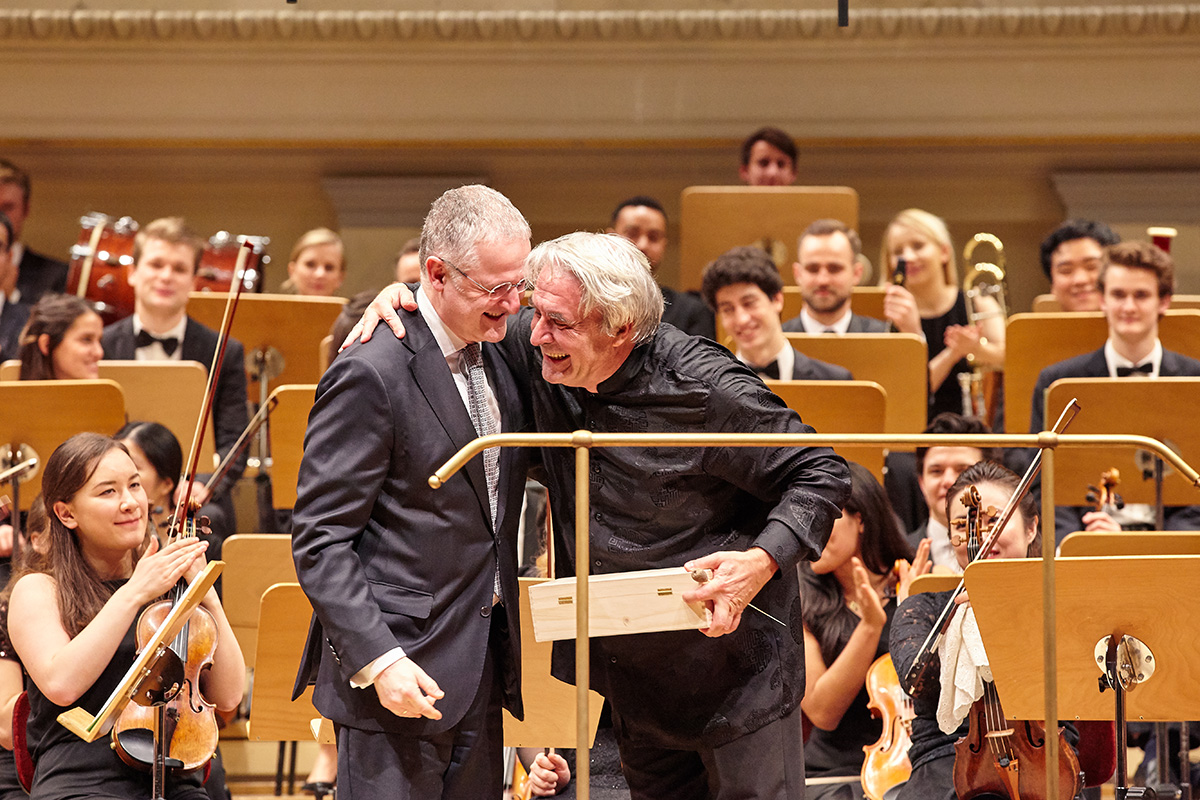 Rektor Robert Ehrlich und Professor Christian Ewald umarmen sich auf der Bühne vor dem Orchester aus Studierenden